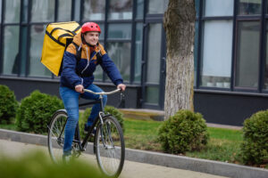 Selective focus of smiling delivery man with thermo backpack riding bicycle on urban street